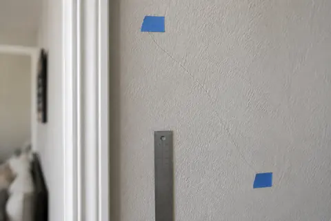 Hairline drywall crack marked with blue painter's tape beside a metal ruler, illustrative image