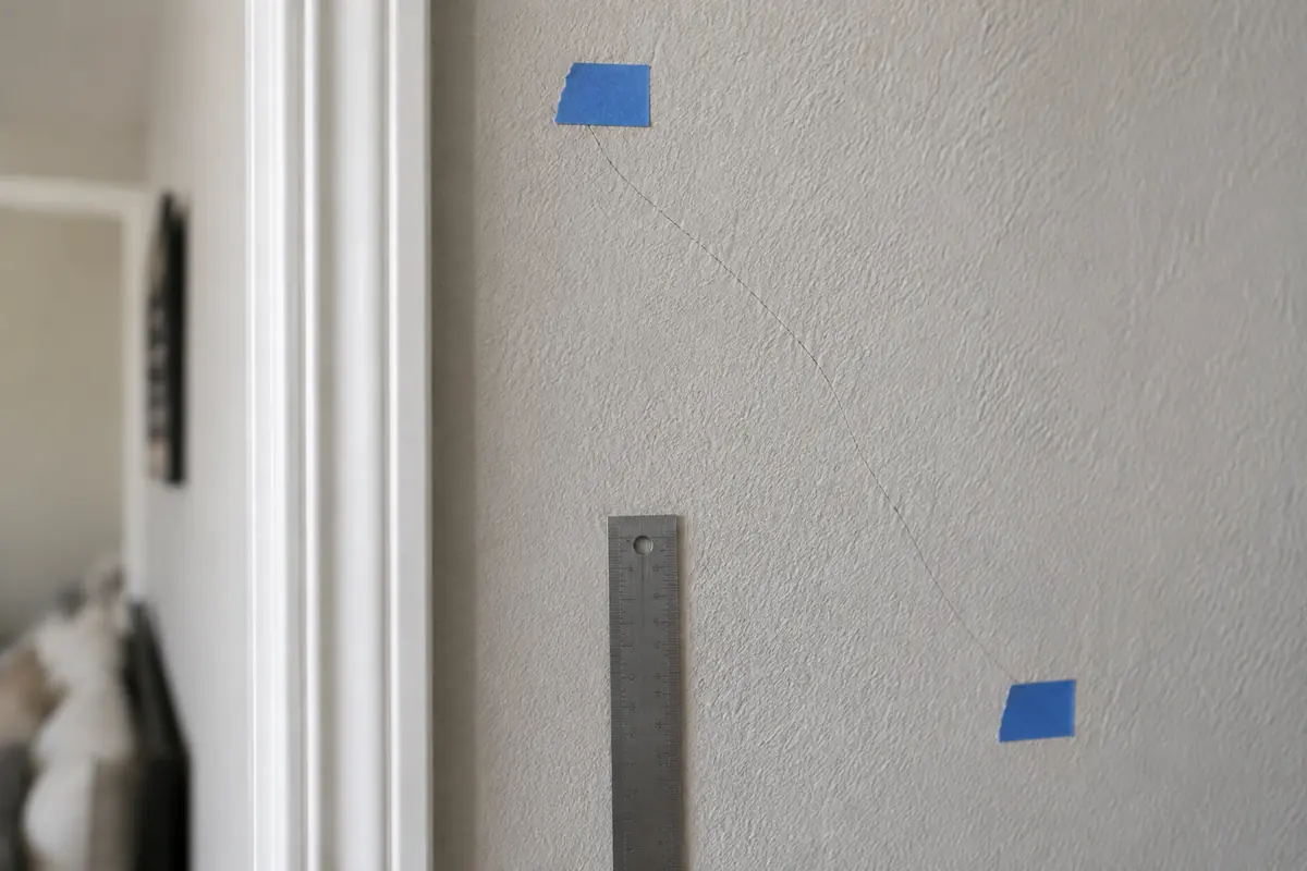 Hairline drywall crack marked with blue painter's tape beside a metal ruler, illustrative image