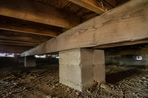 Wooden girder seated on a masonry pier beneath timber floor joists in a crawlspace, illustrative image